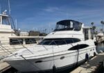 White motor yacht moored at a marina, with a black canvas cockpit cover, stainless rails, and other boats visible in the background.