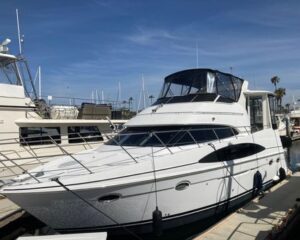 White motor yacht moored at a marina, with a black canvas cockpit cover, stainless rails, and other boats visible in the background.