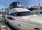 A white motor yacht docked at a marina under a clear blue sky, with a covered upper deck and railing along the bow.