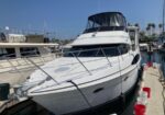 White motor yacht moored at a marina on a sunny day, with a fenced dock, rails, and other boats in the background.