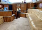 Yacht interior with beige leather sectional, wooden cabinets, and galley area on the left under skylights.