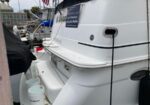 White motor yacht docked at a marina, with a blue sign on the side and mooring lines visible amid nearby boats and gear like buckets and fenders.