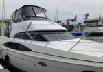 White motor yacht docked at a marina, upper deck covered with a black canvas, with neighboring boats and an American flag in the background.