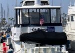 Rear view of a white motor yacht docked in a marina, with a black cover over the aft deck and an American flag fencing the stern.