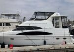 White motor yacht docked at a marina, with a covered cockpit and black window accents; fenders along the hull and other boats visible in the background.