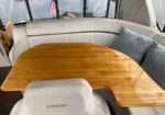 Wooden boat table surrounded by light leather seating and cushions on a marina, with water outside the windows.