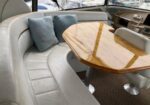 Cozy boat cabin seating area with a curved beige leather sofa, light blue cushions, and a polished wooden oval table on pedestal bases, overlooking a marina.