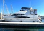 White motor yacht docked at a marina with blue sky and masts in the background.