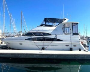 White motor yacht docked at a marina with blue sky and masts in the background.