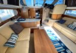 Interior of a motor yacht cabin: beige leather sofas around a wooden coffee table, with a kitchenette and helm area in the background, sunlit windows