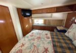 Boat cabin bedroom with a bed, sea-themed quilt, wooden storage cabinets, wall-mounted TV, and a window with beige curtains.