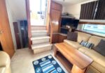 Cozy boat interior with a beige sofa, wooden coffee table, built-in cabinets, and stairs to the upper deck beside a wall-mounted TV.
