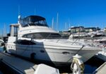 White motor yacht docked at a marina, with other boats in the background under a bright blue sky.