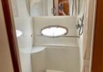 Compact white boat bathroom with a built‑in shower, small porthole window, and curved bench; mirror shows the photographer taking the photo.