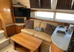 Cozy boat cabin living area with beige sofa, cushions, wooden coffee table, mounted TV, and curved countertop by a window with blinds.