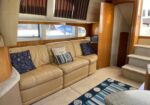 Interior of a yacht living area with a beige leather sofa, blue patterned pillows, wood cabinetry, and a striped rug.