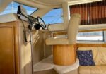 Boat helm in a sunlit cabin with steering wheel, navigation display, throttle, beige captain’s chair, and wood cabinetry.