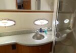 Small yacht bathroom with curved wood vanity, gray speckled countertop, round sink, two porthole windows, and a glass shower stall.