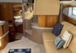 Interior of a boat cabin with a beige leather sofa, helm chair and control panel, wooden cabinetry, and stair to the wheelhouse; blue striped rug on light carpet and windows with brown curtains.