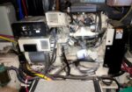 Close-up of a diesel generator engine with belts, hoses, and a control box in a cramped mechanical cabinet.