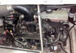 Close-up of an engine bay with black hoses, metal pipes, wiring, and a translucent coolant reservoir in the background.