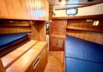Wood-paneled boat cabin with two blue bunks, storage cabinets, and a small desk area seen through a central doorway.