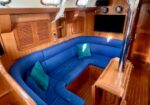 Wood-paneled boat cabin with a curved blue sofa, teal pillows, a small wooden table, and a wall-mounted TV.