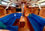Wood-paneled sailboat cabin with blue U-shaped seating, a central wooden table, and a metal mast post; portholes and hatches along the walls create a cozy living area.
