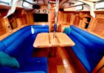 Interior of a sailboat cabin: U-shaped blue cushioned seating around a wooden table, with a central pole, teak walls and floor, and built-in storage.