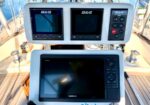 Boat helm showing three B&G navigation displays above a Garmin chartplotter; sailing instrument cluster and controls on a teak deck.