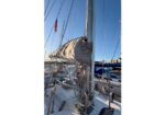 Sailboat deck with a folded mainsail cover on the boom, rigging and lines, mast, and a marina with other boats and an American flag nearby.