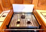 Three-burner stainless steel gas stove with a white hinged lid, built into a wooden boat galley with varnished panels on either side