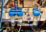 Under-sink water filtration system with three blue-filter housings and clear cartridges connected by blue tubing.