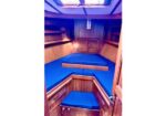 Wood-paneled boat cabin with blue cushioned seating arranged in a V shape around a central step, with a skylight overhead.