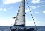 Sailing yacht with white sails on a calm blue ocean, American flag at the stern under a clear sky.