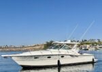 White luxury motor yacht with two tall antennae anchored on calm blue water, harbor and buildings in the background against a clear sky.