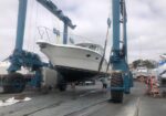 A white boat is being lifted out of the water by a large blue travel lift at a dry dock marina.