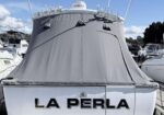 Rear view of a white yacht with a gray canvas cover over the upper deck, docked in a marina; the boat is named 'LA PERLA' on the stern.
