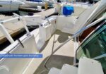White boat cockpit with an open transom gate for boarding, docked at a marina; blue cooler cover and neighboring boats in the background.