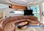 Curved brown leather sofa in a yacht salon with a round wood coffee table, wall-mounted TV, light wood cabinetry, and large windows showing daytime exterior.