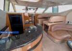 Luxury yacht interior: port-side galley with curved granite counter, wooden cabinets, fridge, and beige leather seating by large windows.