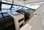 Yacht cockpit and helm with white seating, wood trim, steering wheel, instrument panel, and large windshield; docked boats visible outside.