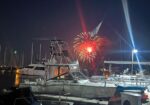 Night-time marina with several yachts docked; red fireworks bloom above the water and reflect on the boats and calm harbor.