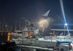Night scene at a marina with boats docked and fireworks lighting the sky.
