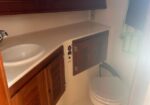 Small bathroom with white sink and wood vanity, an open access panel beside it, and a closed toilet; a green towel hangs by a sunlit window.