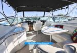 Interior of a motor yacht's flybridge: white cushioned seating, helm with steering wheel, curved table, and a canopy, with marina visible through windows.