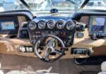 Luxury boat cockpit dashboard featuring a central steering wheel, multiple analog gauges, two navigation screens on either side, beige leather panels, and various control switches; marina and other boats visible through the windshield.