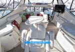 Luxe yacht flybridge seating area with white leather U-shaped sofa, a central white table, colorful cushions, and a mounted TV, overlooking a marina.