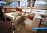 Cream leather yacht salon with U-shaped seating around a wood table and decorative cushions, windows with blinds in view.