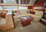 Cozy beige leather sectional sofa with colorful cushions in a yacht’s lounge, wooden coffee table with magazines, and wood-paneled cabinets by large windows with blinds.